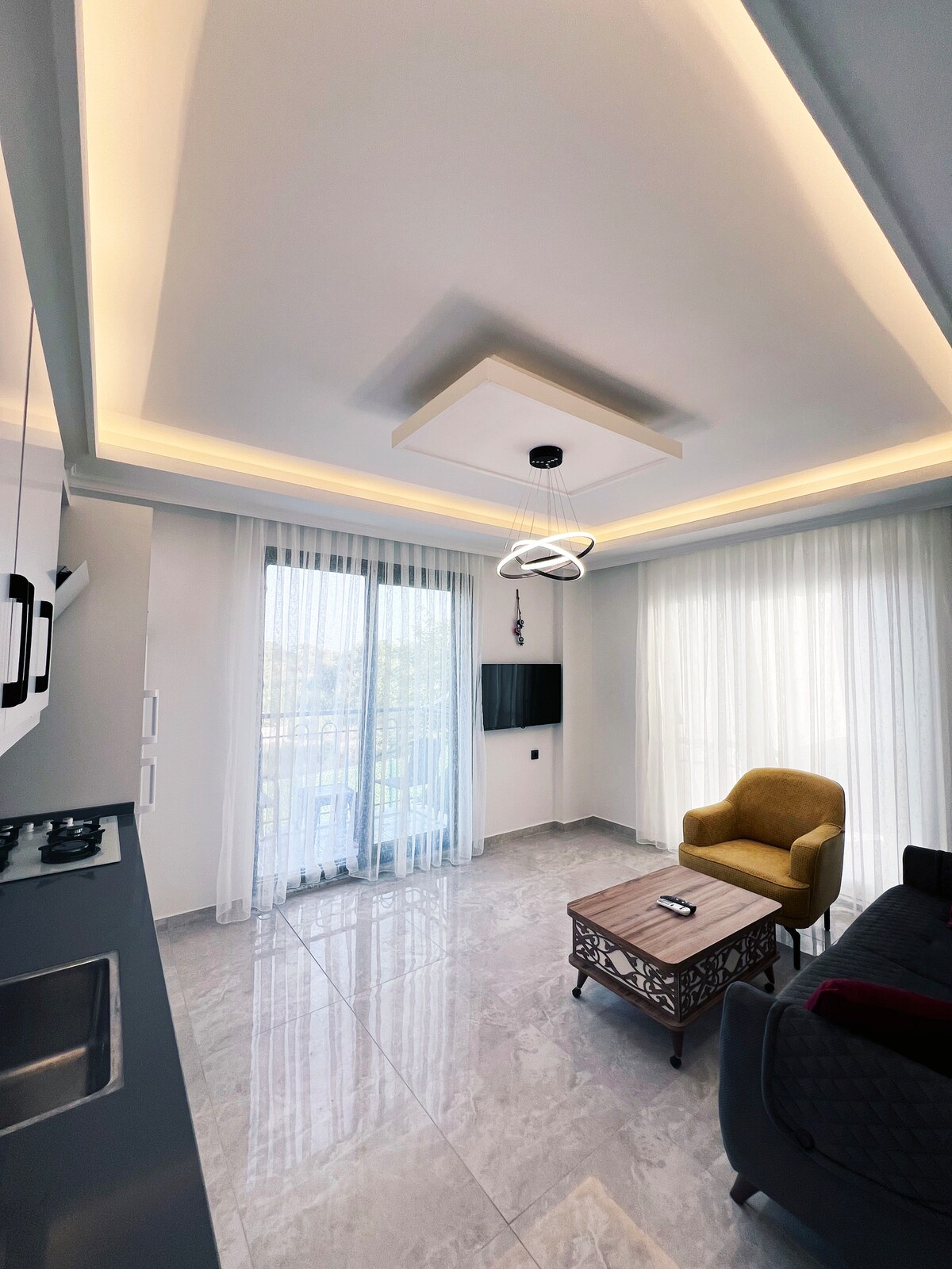 A modern living space features a sleek design with a high ceiling illuminated by recessed lighting. Large floor-to-ceiling windows draped in light sheer curtains allow natural light to fill the room. A comfortable yellow chair and an elegant coffee table complement the cozy atmosphere.
