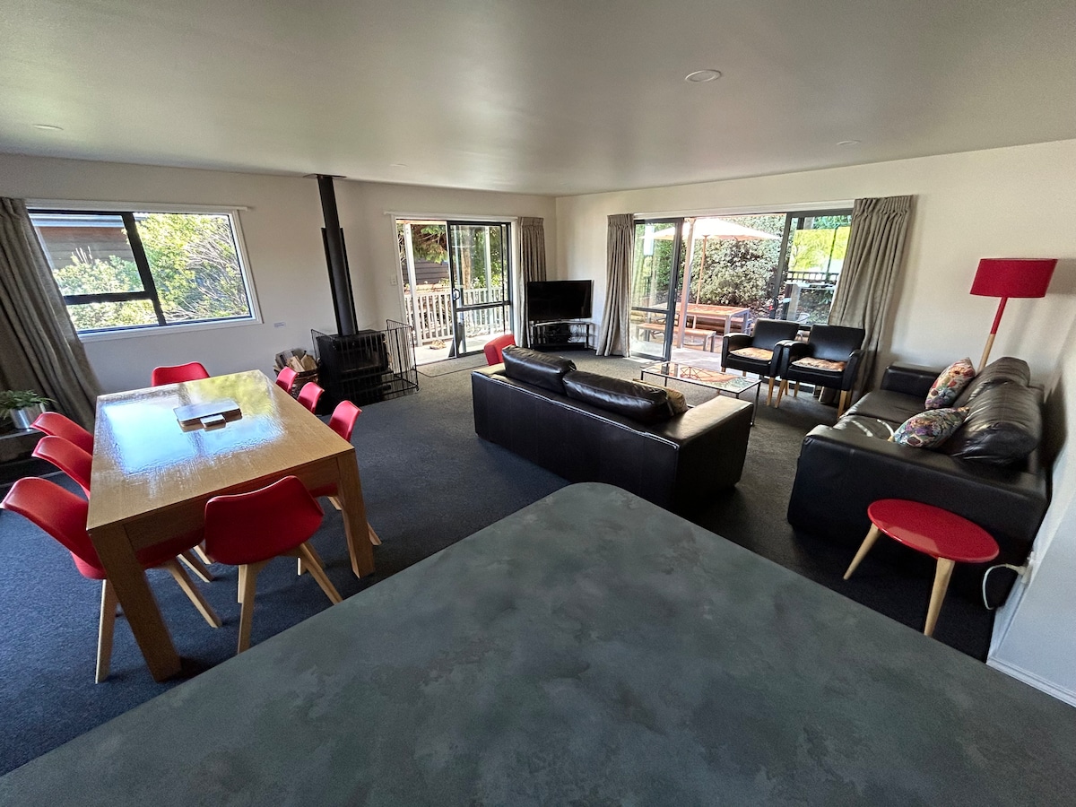 An open-plan living area features large windows allowing natural light. A dining table with red chairs is positioned near the window, while two sofas and a TV provide a comfortable seating arrangement. A log fire enhances the cozy vibe, with sliding doors leading to the patio.