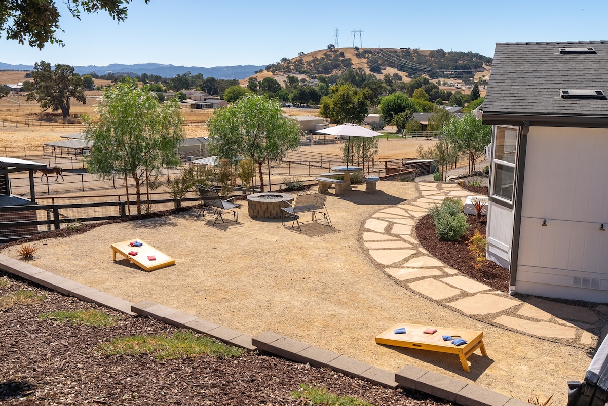 An outdoor area features a pathway winding through a landscaped yard with seating arrangements around a fire pit. Two cornhole boards are positioned on the ground, while lush trees provide shade. The backdrop showcases rolling hills and open fields under a clear blue sky.