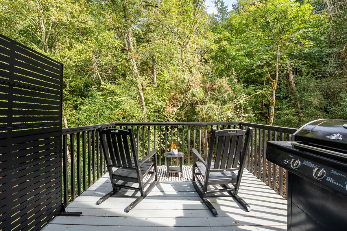 A private deck features two black rocking chairs and a side table, overlooking a natural landscape filled with lush trees. A propane BBQ grill is positioned nearby, contributing to a serene outdoor space.