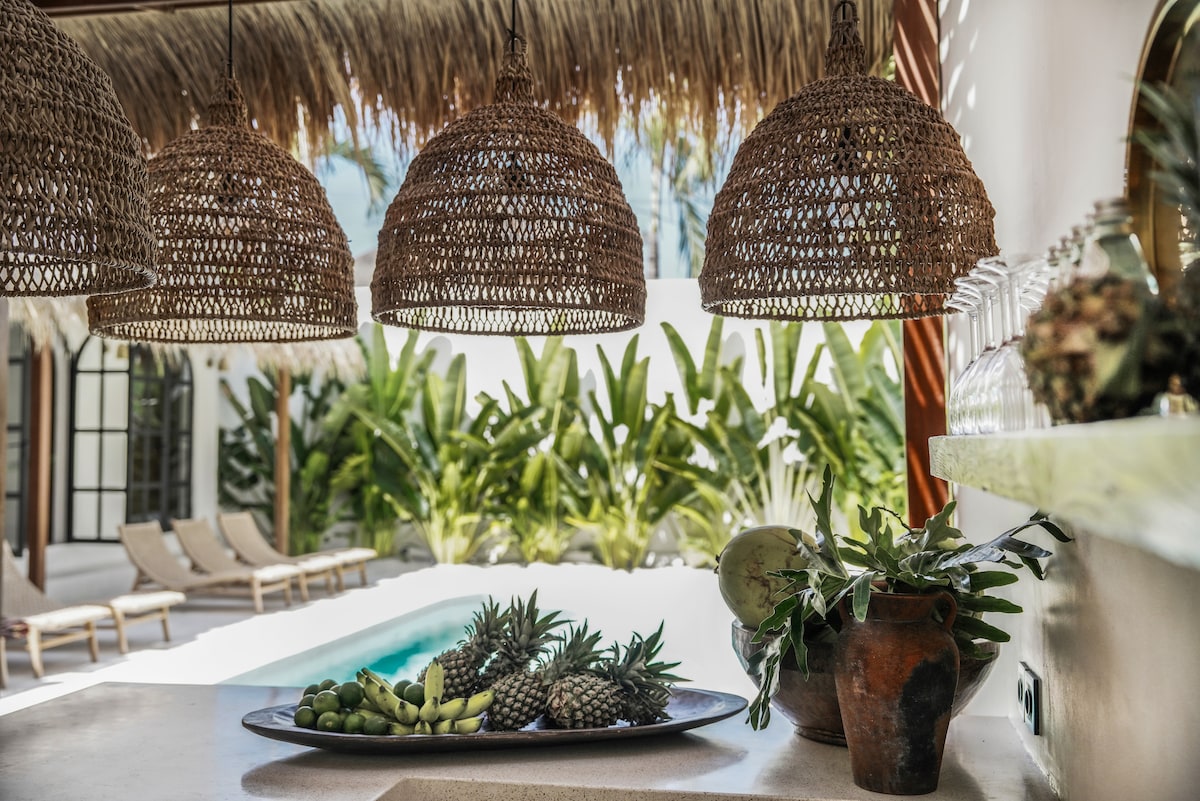 A kitchen bar area features woven pendant lights above a countertop adorned with a decorative tray. The tray displays a selection of fresh fruits, including pineapples and limes. Beyond the countertop, a tranquil pool and lush greenery are visible, enhancing the inviting atmosphere.