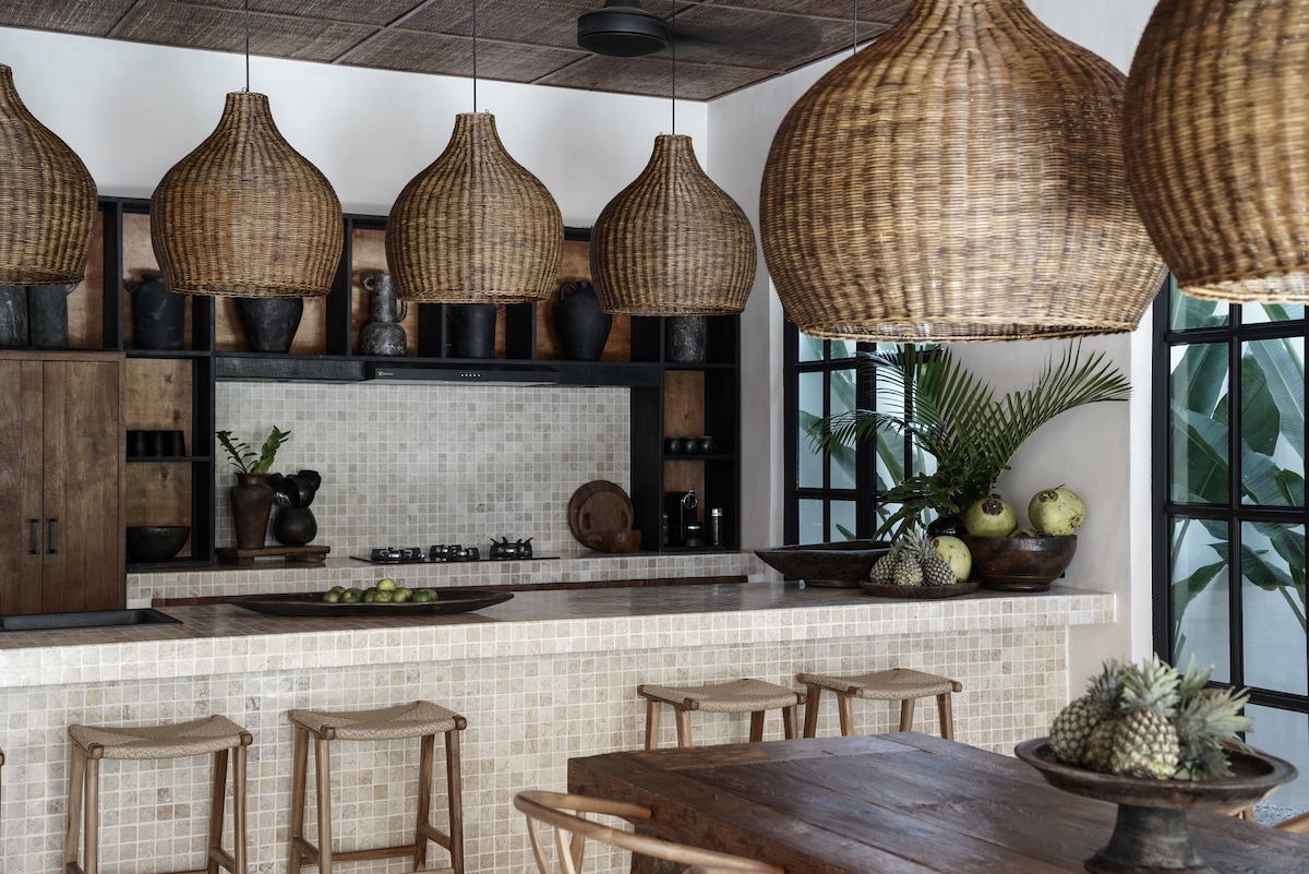 A fully equipped kitchen features unique marble mosaic countertops and a kitchen island with seating. Rattan pendant lights hang above, complementing the natural decor with dark ceramic accents and plants. Floor-to-ceiling windows provide views of greenery, enhancing the space's connection to nature.