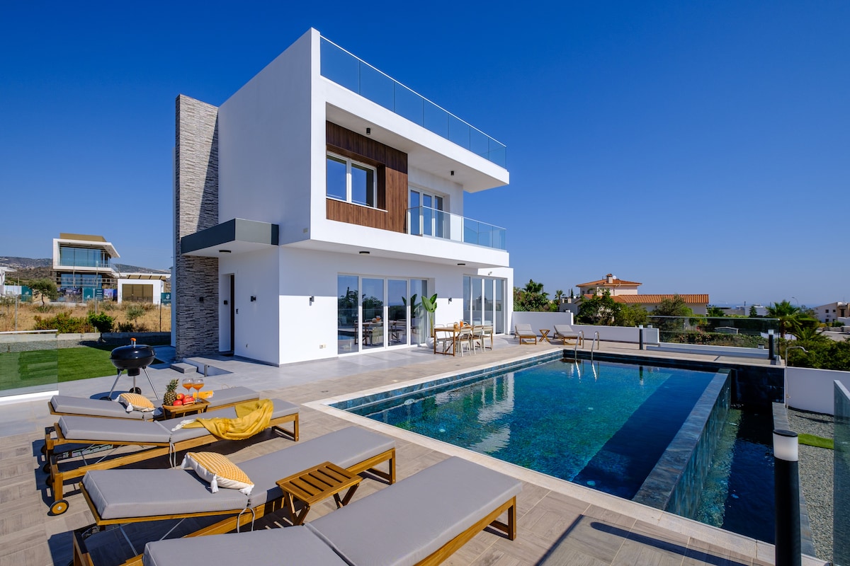 The modern villa stands elegantly with a spacious outdoor area featuring an infinity pool. Sun loungers are arranged around the pool, providing a comfortable space for relaxation. Large glass doors lead into the villa, emphasizing the bright and open design.