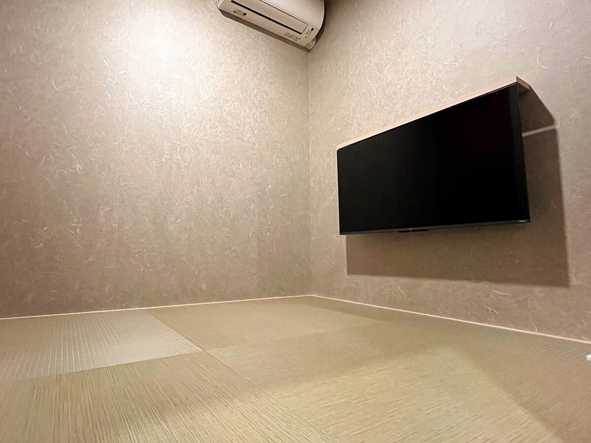 A flat-screen television is mounted on the wall in this compact room featuring tatami mat flooring. Neutral-colored walls and air conditioning create a simple yet comfortable environment for relaxation or entertainment.