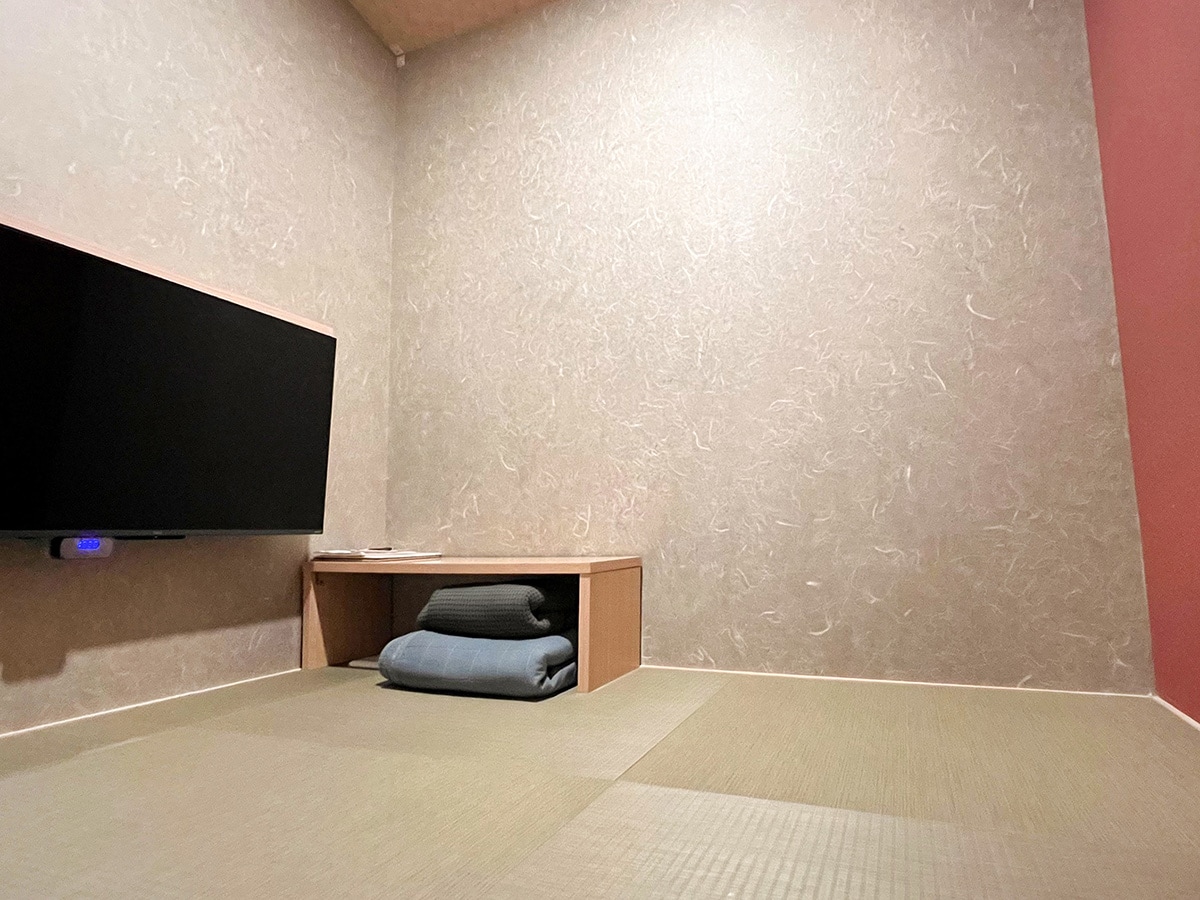 A minimalist room features light-colored walls with a smooth texture, complemented by soft tatami flooring. An LED television is mounted on the wall, and a low wooden table sits adjacent to neatly folded bedding items.