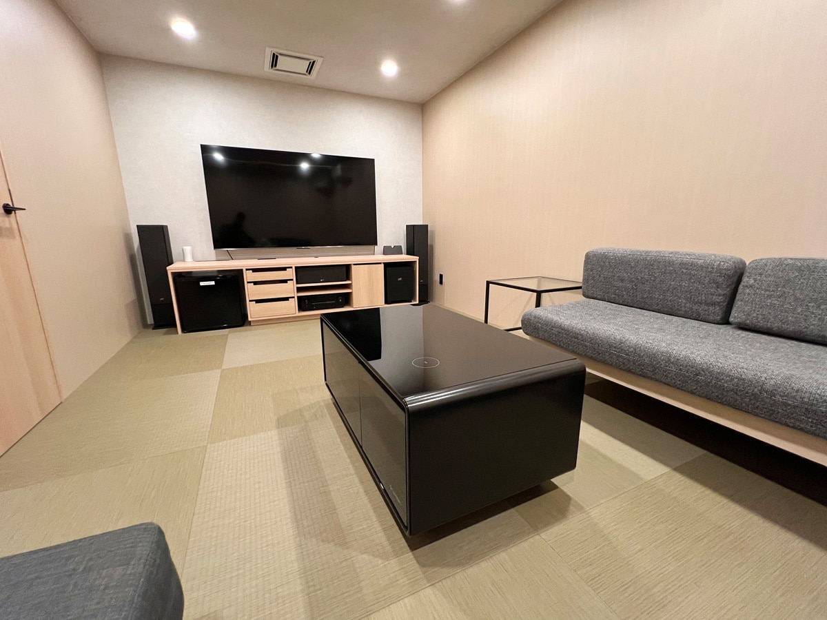 A modern theater room features a large flat-screen television mounted on the wall, complemented by a light wood entertainment unit. A dark coffee table is centered among the seating area, which includes a gray sofa and a smaller side table, all set on muted tatami flooring.