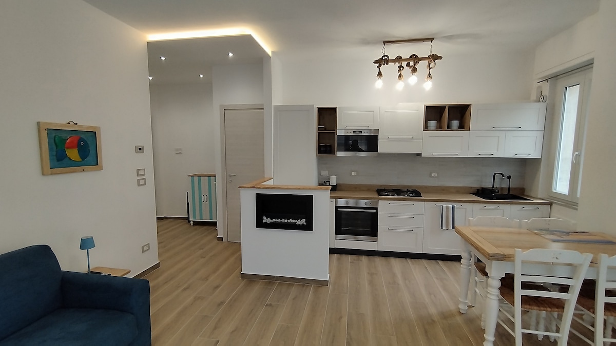 A modern kitchen is visible, featuring white cabinetry and stainless-steel appliances. A wooden countertop and a small dining table with white chairs provide a cozy dining area. Light-colored wood flooring creates a warm ambiance, complemented by natural light coming through the large windows.