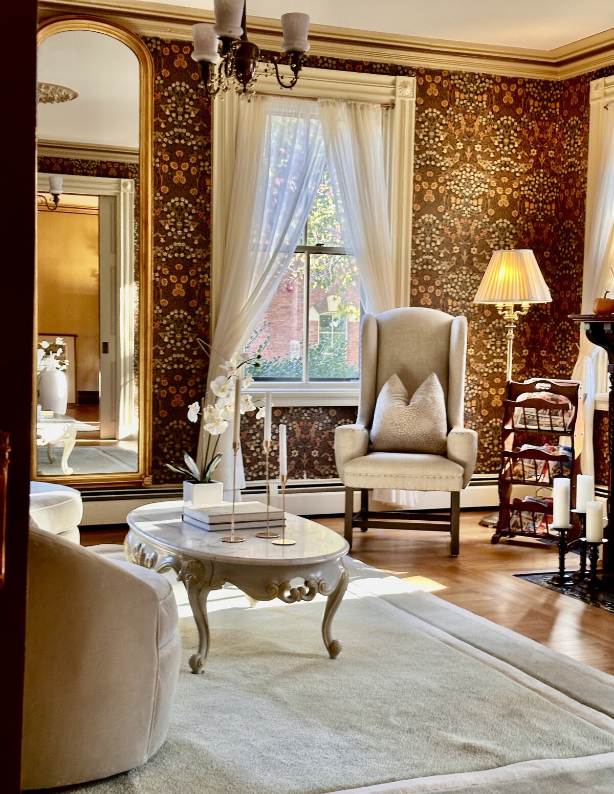 A formal living room is adorned with floral wallpaper and natural light streaming through the window. A plush armchair stands next to a delicate coffee table, with elegant decor and candles arranged on it. A large mirror reflects the tranquil scene, enhancing the room's spacious feel.
