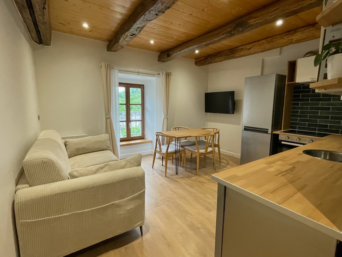 A cozy living space features a comfortable couch and a wooden dining table surrounded by chairs. Natural light filters through a window, with views of greenery outside. The kitchen area is equipped with modern appliances, and exposed wooden beams add character to the ceiling.