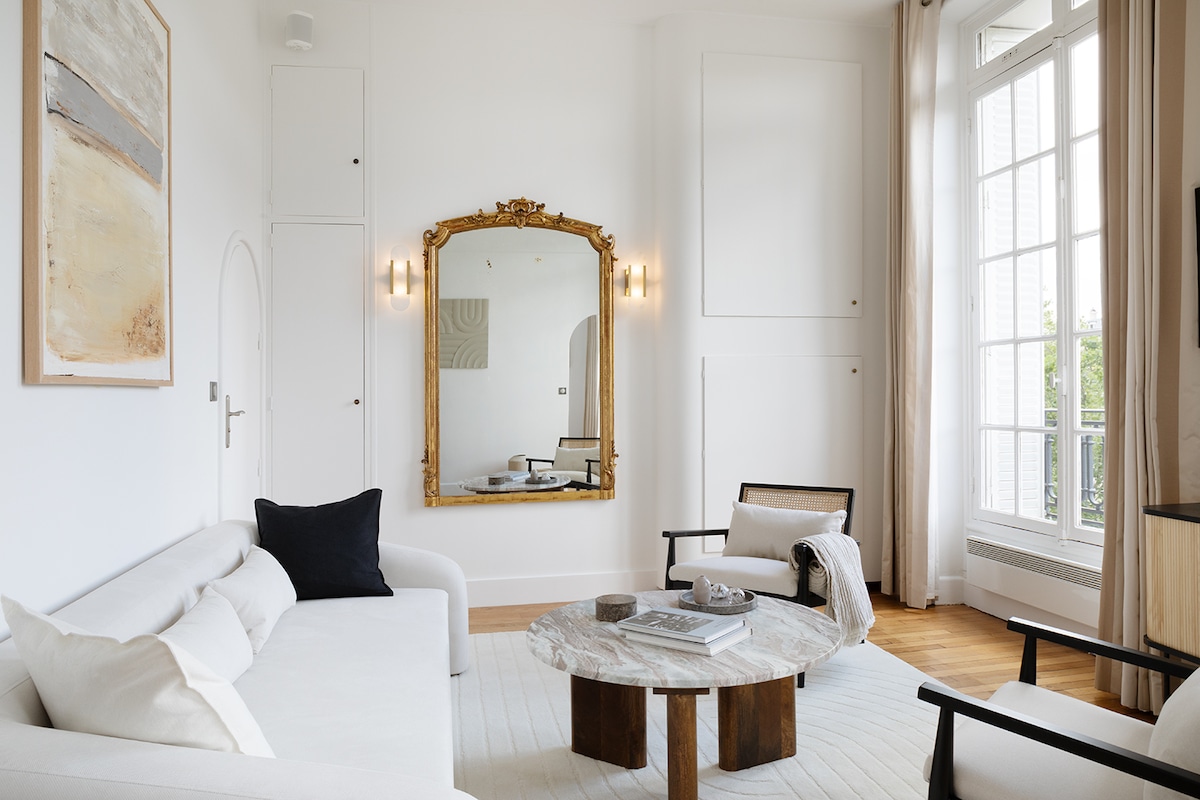 A spacious living room is highlighted by a large mirror framed in gold, reflecting the light-filled space. A comfortable sofa with decorative cushions faces a stylish coffee table. Elegant artwork adorns the wall, complementing the minimalist decor and large windows that offer a view of the outside.