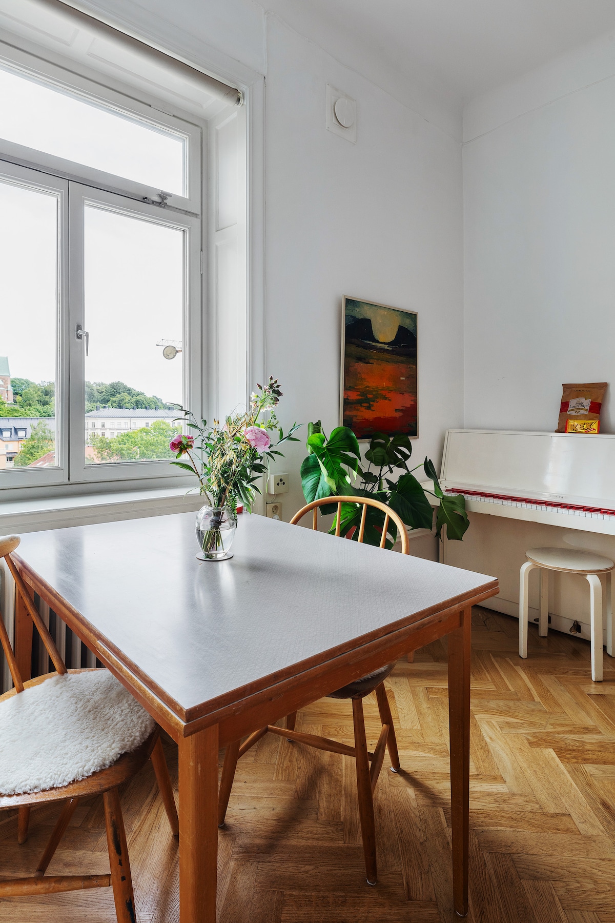 A dining area features a wooden table paired with simple chairs. A vase of fresh flowers sits at the center, while a piano stands in the corner, accompanied by a small stool. Natural light streams in through the window, illuminating the space.