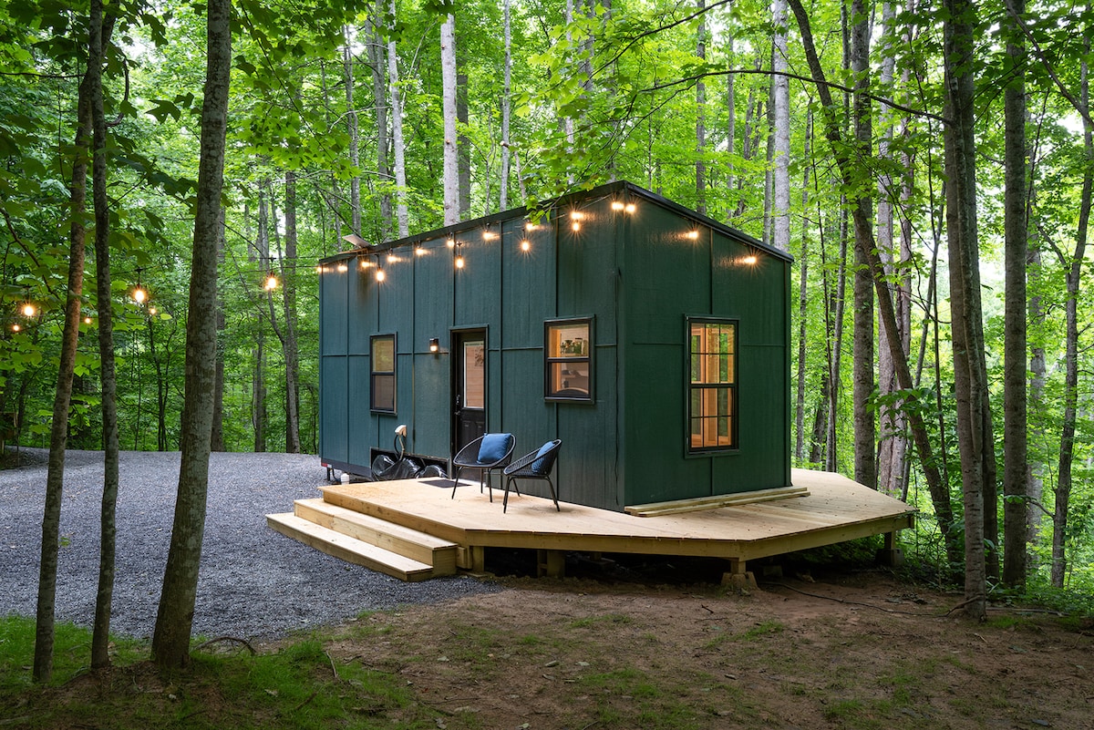 A modern green cabin is nestled among tall trees, featuring a spacious deck with outdoor seating. Soft lighting strung above enhances the natural setting. The gravel path leads to the entrance, while large windows provide glimpses of the tranquil forest surroundings.