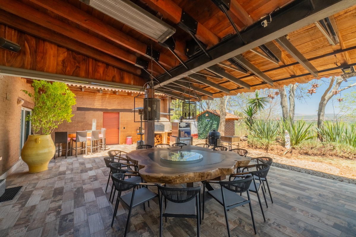 An outdoor dining area features a rustic wooden table surrounded by black chairs under a wooden roof. Sunlight filters through the open space, illuminating the setting. A large clay pot and barbecue station can be seen in the background, enriching the inviting atmosphere.