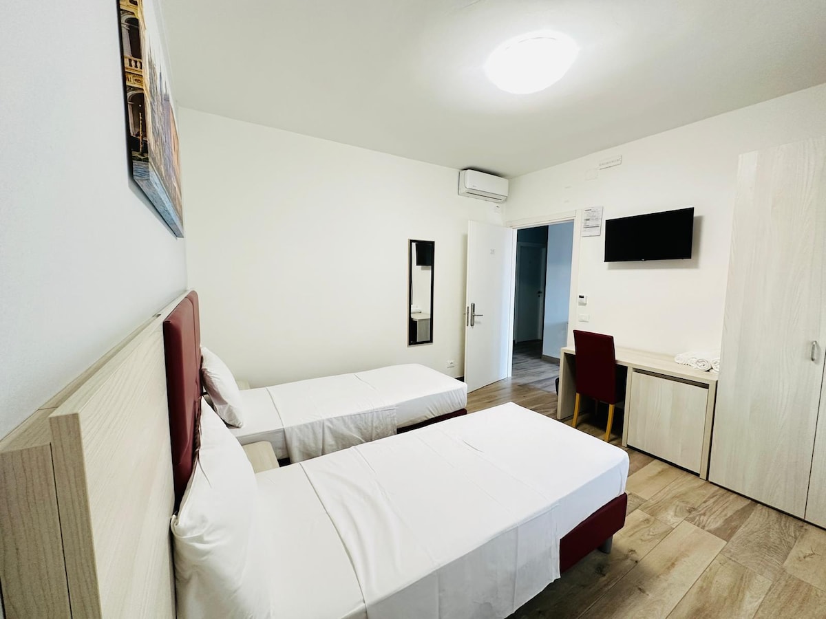 A simple and functional room is designed with two single beds dressed in white linens. A wooden desk and chair are positioned against the wall. A flat-screen television is mounted above the desk, while natural light is provided through a window, enhancing the overall brightness of the space.