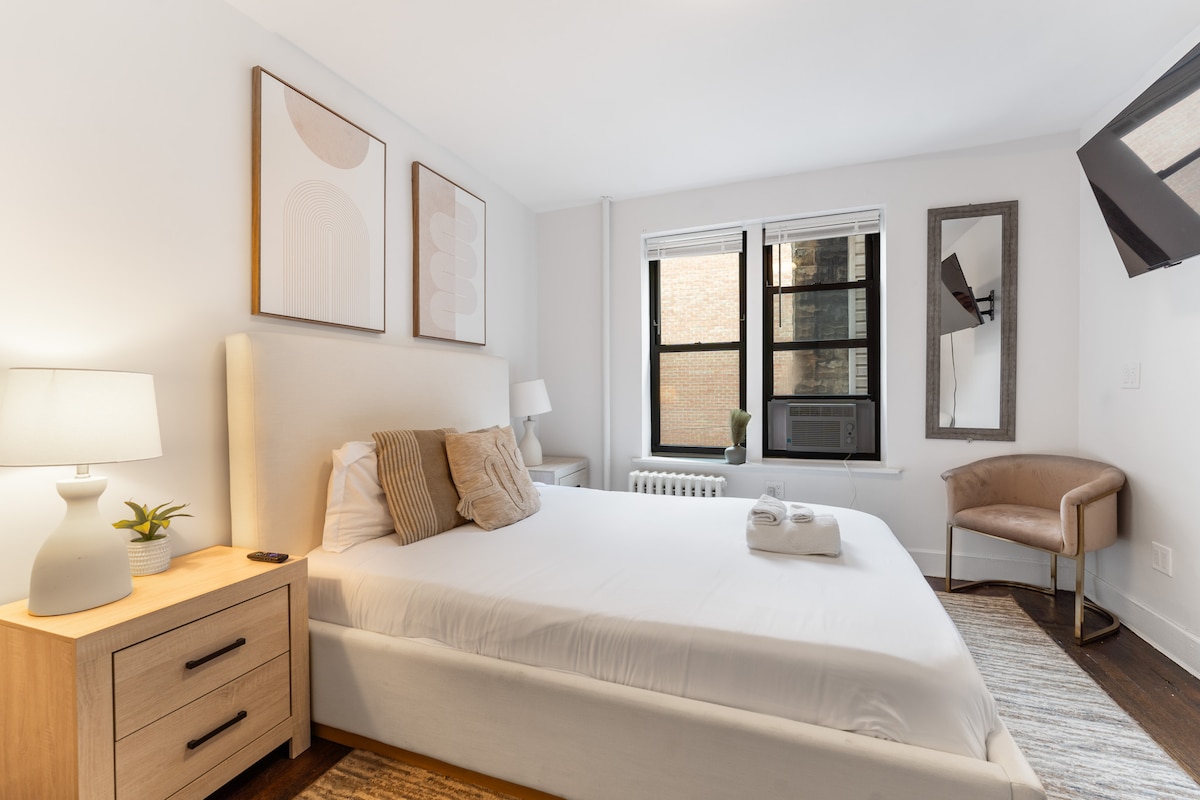 The studio features a spacious bed with crisp linens, complemented by decorative pillows. A bedside lamp and a potted plant are visible on a light wood nightstand. Two windows allow natural light into the room, while a wall-mounted television and an accent chair add functionality to the space.