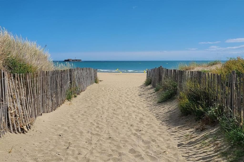 A sandy pathway, bordered by wooden fencing and tufts of grass, leads to a serene beach. The view opens to the azure sea, with gentle waves lapping at the shore. A clear blue sky stretches overhead, contributing to a tranquil coastal atmosphere.