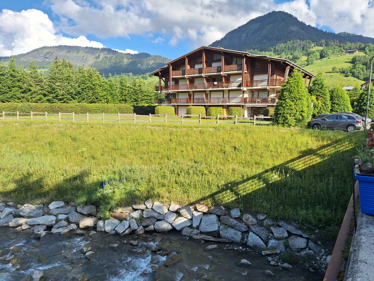 A charming building is set against a backdrop of rolling hills and mountains. The structure features multiple balconies surrounded by lush greenery and a tranquil stream at the foreground, offering a serene outdoor environment.