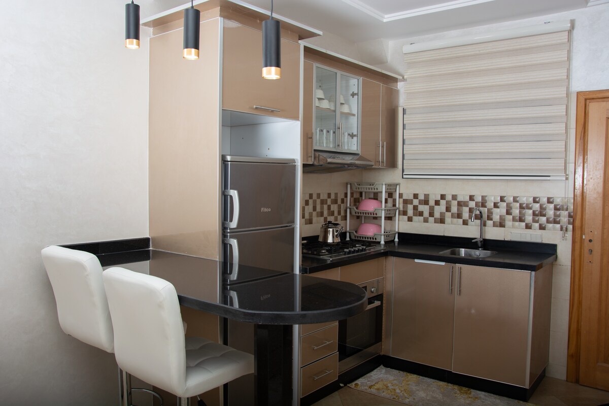 The kitchen features modern cabinetry with a sleek countertop and a matching backsplash. Stainless steel appliances include a refrigerator and an oven. A small breakfast bar with two white stools provides a casual dining option. Natural light enters through a window adorned with light shades.