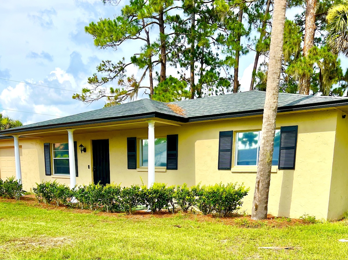 The exterior of the bungalow features a light yellow façade with dark shutters. A covered porch is accessible from the front, surrounded by manicured greenery. Tall trees are visible in the background, and the sky is partly cloudy.