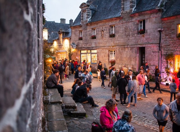 A lively village street scene is captured in the early evening, with stone buildings showcasing their character. Groups of people gather and converse, while window boxes filled with flowers add charm. The cobblestone pathway suggests a warm community atmosphere, illuminated softly by light from nearby shops.