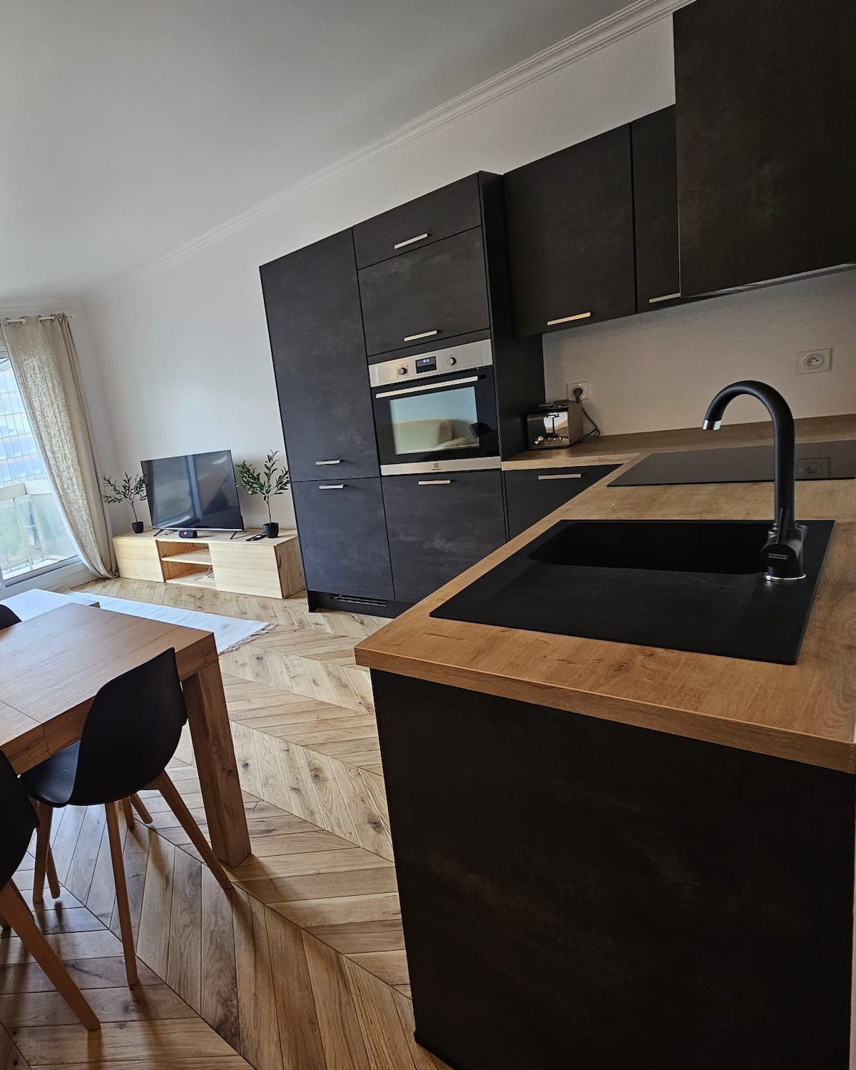 A modern kitchen is viewed with sleek black cabinetry and wooden accents. An integrated oven and stovetop are positioned among the storage units. A dining table, accompanied by black chairs, is visible, complemented by a light-colored rug and a television in an adjacent area.