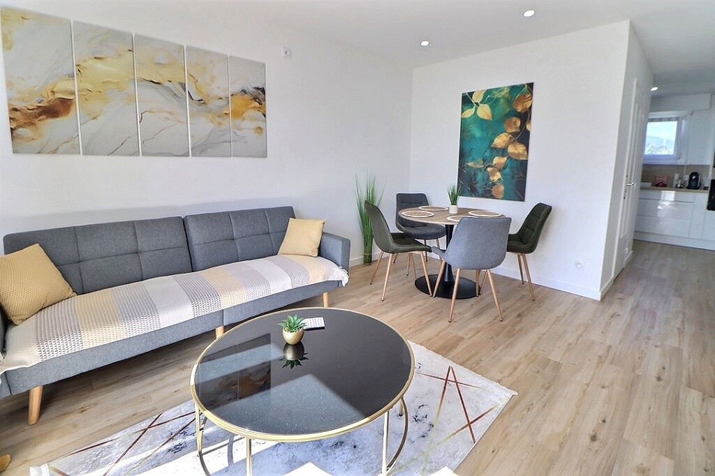 A light-filled living area is presented with a gray sectional sofa and a round dining table set for four. Abstract artwork adorns the walls, while indoor plants add a touch of greenery. The open layout leads to a modern kitchen visible in the background.