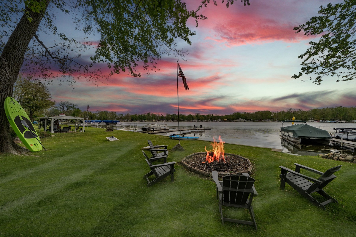 A fire pit surrounded by several chairs is set on a grassy area, providing a cozy gathering spot. A lake view is visible in the background, with docks and boats. A vibrant sunset sky adds color, while a kayak is propped against a tree nearby.