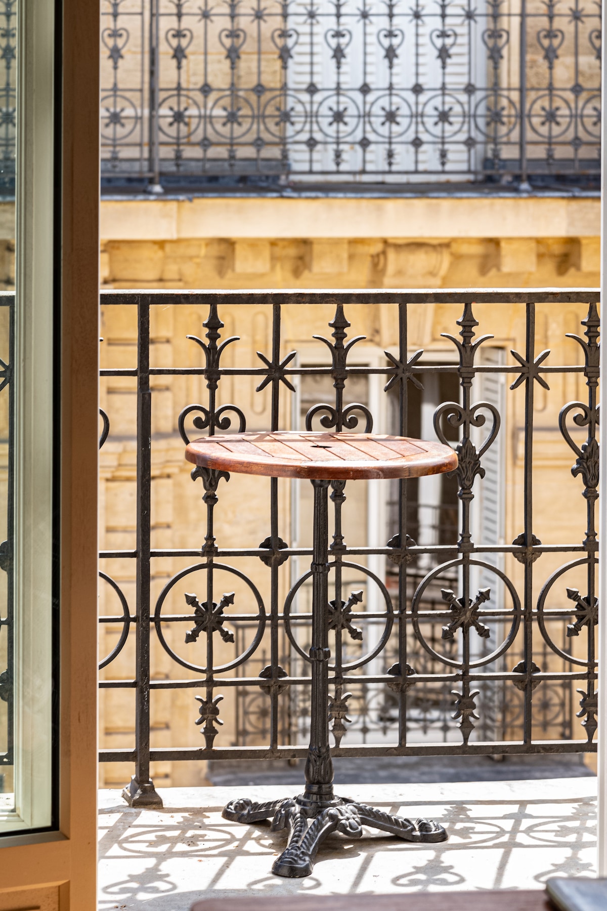 A small, round table is positioned on a wrought iron balcony with intricate patterns. The balcony railing features decorative scrollwork, while soft light illuminates the warm tones of the building in the background, enhancing the outdoor space.