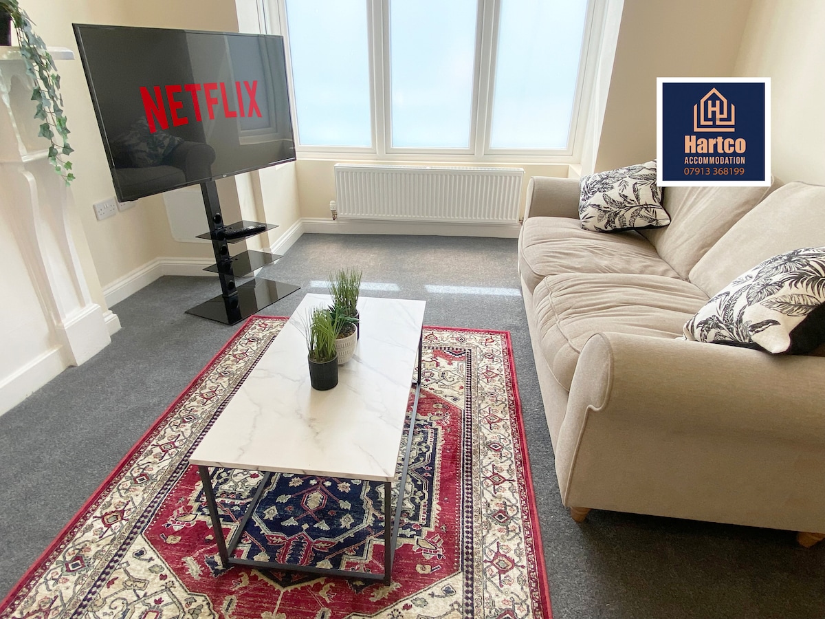A comfortable living area is featured with a neutral sofa positioned beside a marble-top coffee table. A large TV displaying Netflix is visible, and natural light streams through the windows, illuminating the soft gray carpet and decorative rug beneath.