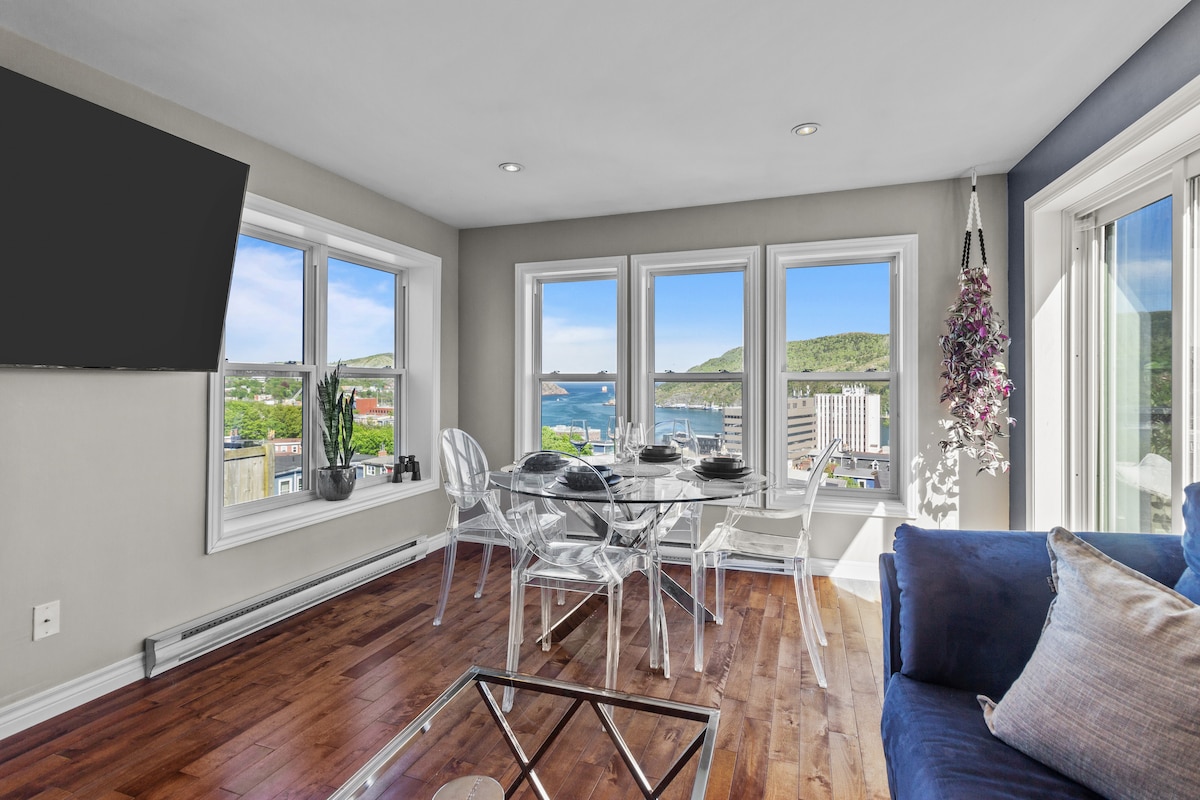 A light-filled living area features large windows offering views of the harbor and mountains. A glass dining table, set for four, complements clear acrylic chairs. A plush blue couch with decorative pillows provides a cozy seating option, while a mounted TV is positioned nearby.