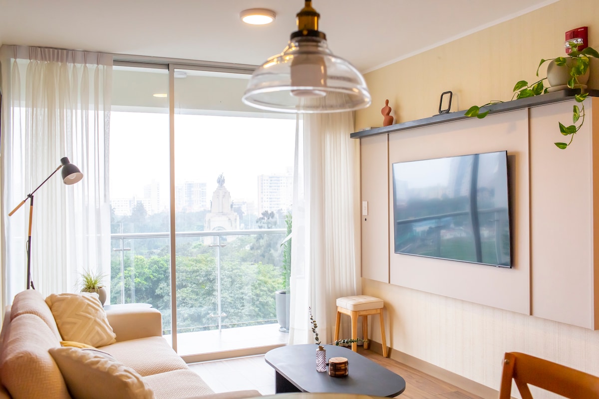 A bright living area features a light-colored sofa paired with a minimalist coffee table. A large flat-screen television is mounted on the wall, while floor-to-ceiling windows provide views of the surrounding greenery. Soft curtains gently diffuse the natural light entering the space.