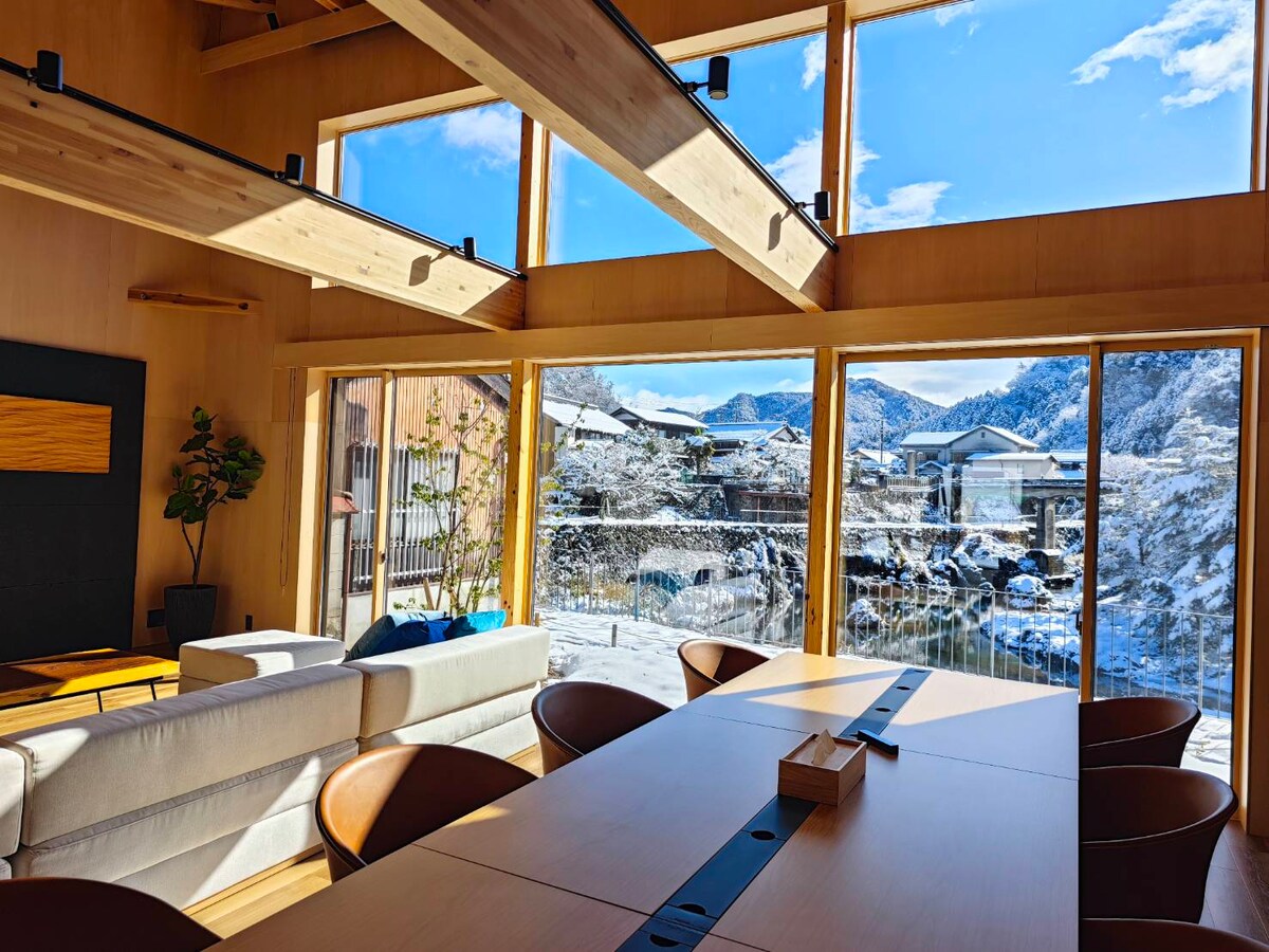 A modern dining area features a large wooden table surrounded by sleek chairs, illuminated by ample natural light through expansive windows. The view reveals a snow-covered landscape, with mountains and traditional houses in the background, creating a serene atmosphere.