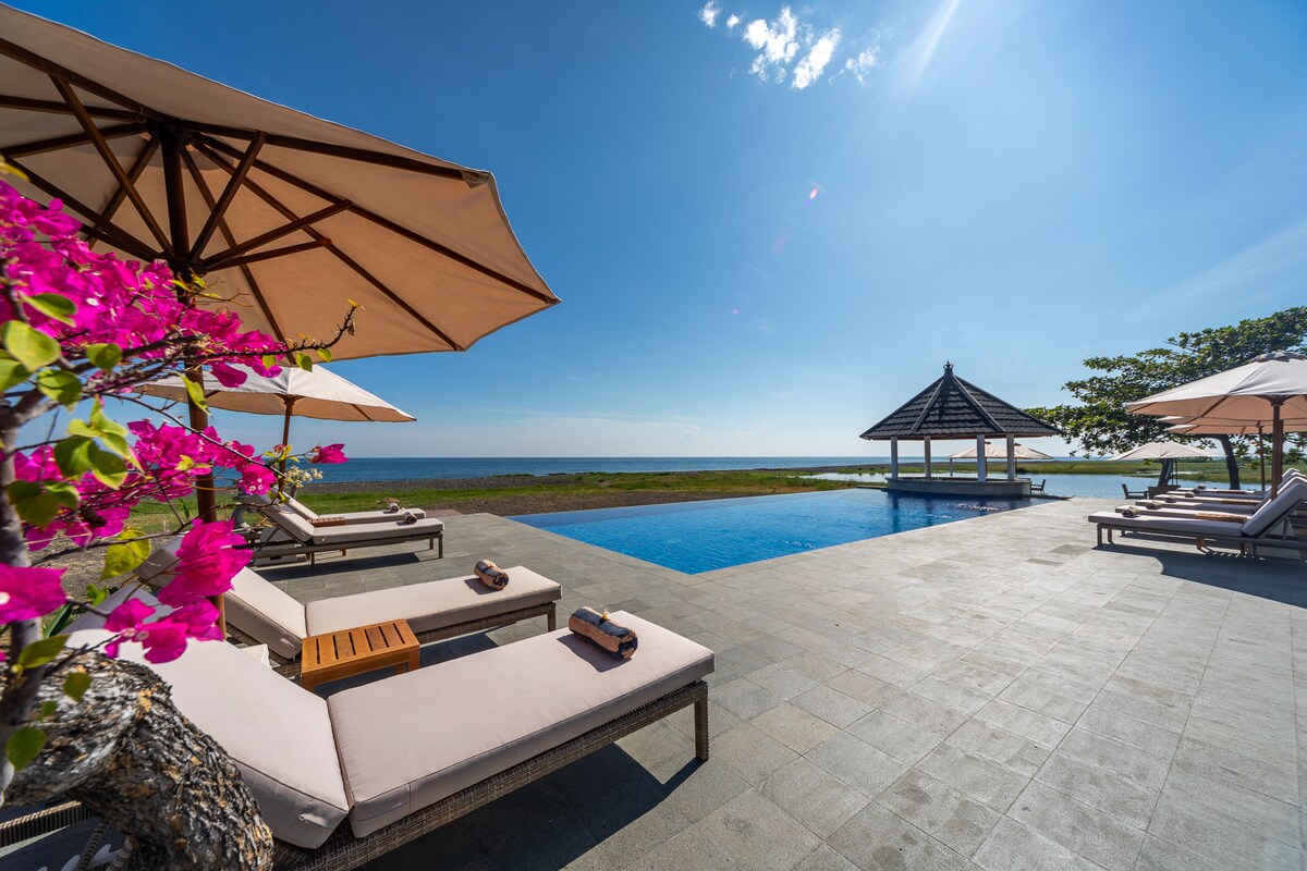 A serene outdoor area features a clear blue sky and a tranquil pool. Lounge chairs are arranged near the water, accompanied by umbrellas providing shade. Vibrant pink flowers add a pop of color to the scene, enhancing the calm ambiance surrounding the space.