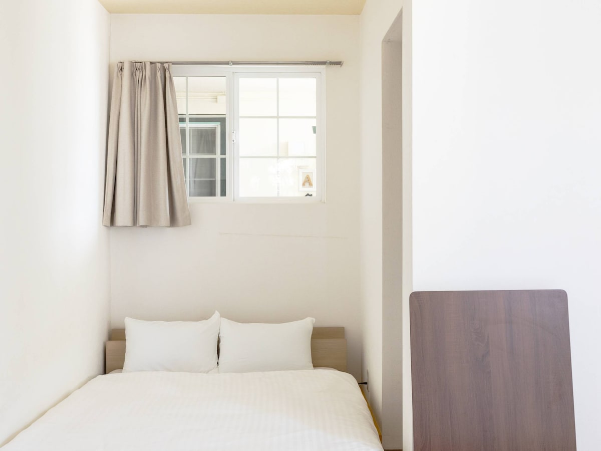 A comfortable sleeping area features a spacious bed with two white pillows, complemented by light-colored bedding. A window with beige curtains allows natural light to enter, while a wooden table stands nearby against the wall.