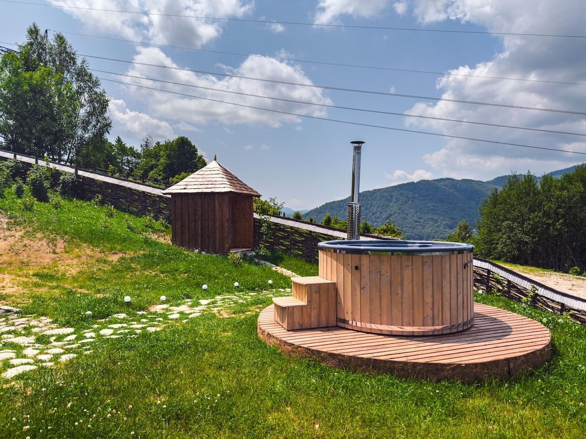 An outdoor hot tub is set on a wooden deck surrounded by lush green grass. A nearby wooden structure features a traditional design with a conical roof. Scenic mountain views and a clear sky create a serene atmosphere.