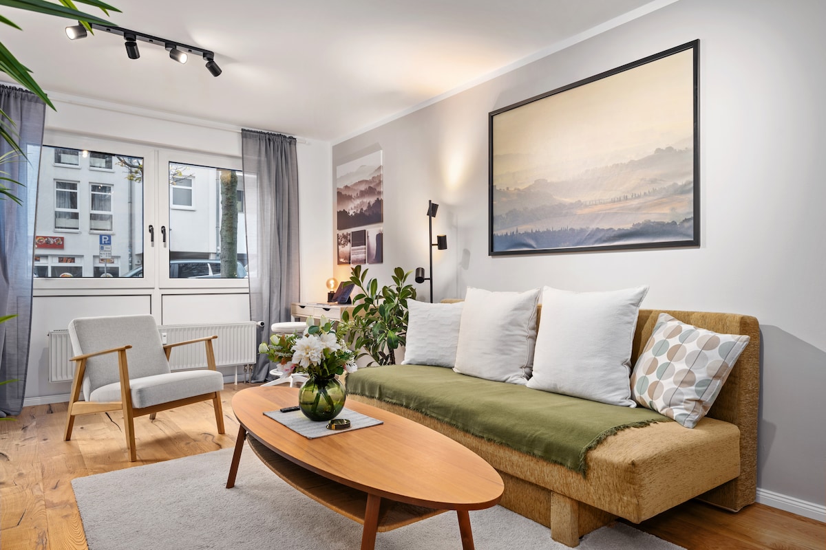 The spacious living room features a cozy sofa adorned with decorative cushions and a soft green throw. A sleek wooden coffee table rests on a light-colored rug, complemented by a potted plant. Large windows enhance natural light, framed by gray curtains.