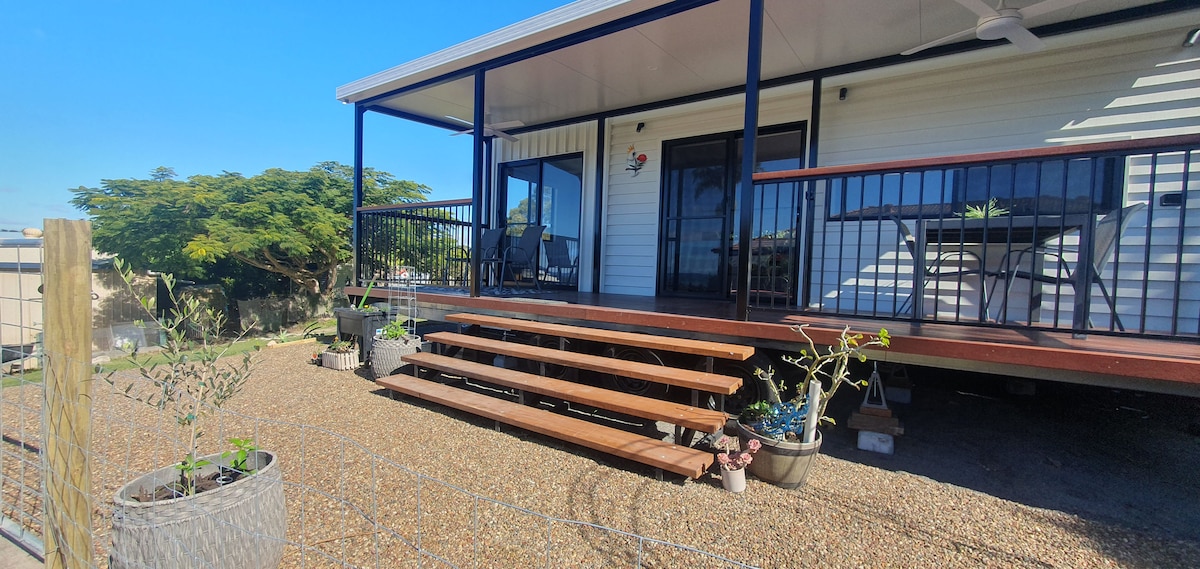 A welcoming porch with wooden steps leads to a tiny home, framed by potted plants on either side. The outdoor seating area features a small table and chairs, while the home displays large windows and a covered roof, offering a shaded space for relaxation.