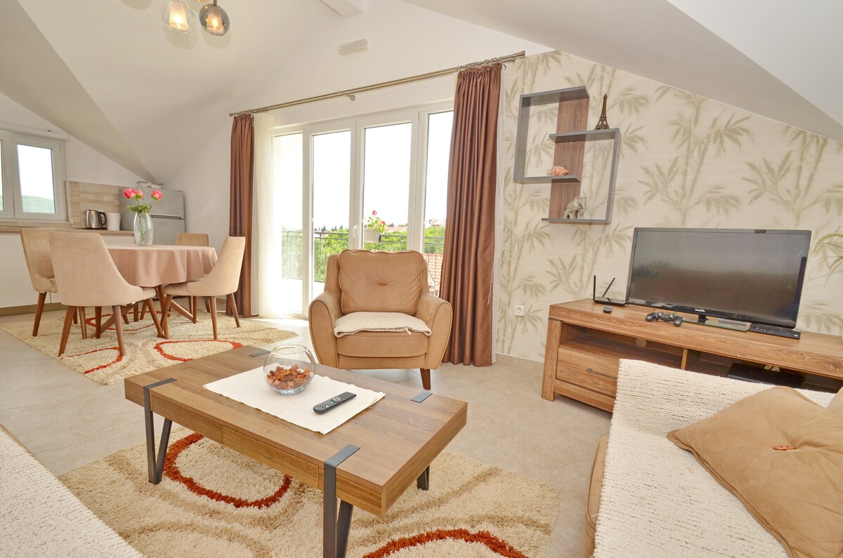 An inviting living space features a comfortable sofa and wooden coffee table, accompanied by a small decorative bowl. A TV is positioned on a wooden stand. The dining area includes a round table surrounded by four chairs, with large windows offering scenic views. Soft lighting enhances the ambiance.