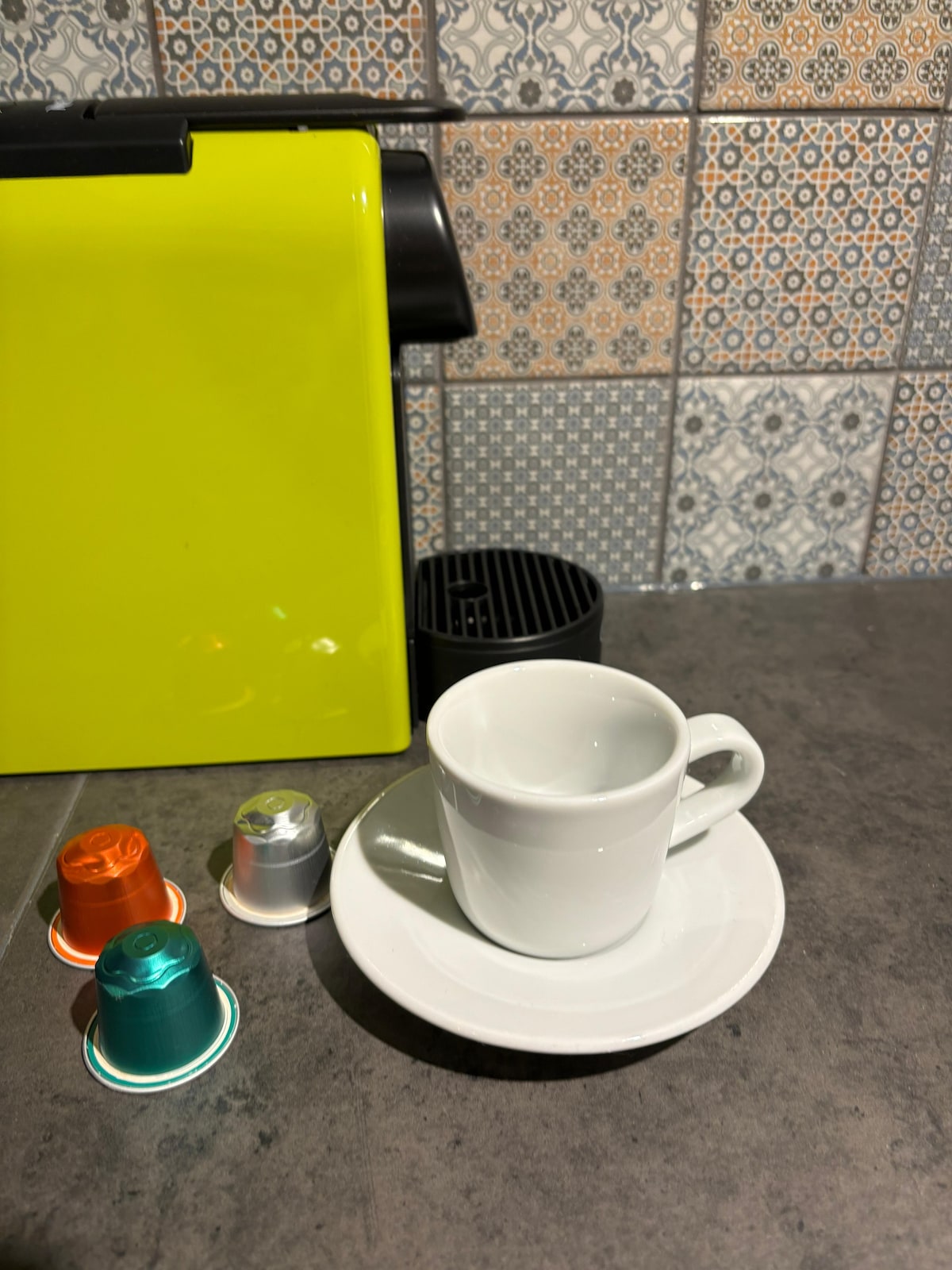 A vibrant yellow Nespresso coffee machine is positioned beside a small white espresso cup and saucer. Several colorful coffee capsules in orange, silver, and teal are arranged nearby, against a backdrop of patterned tiled wall.