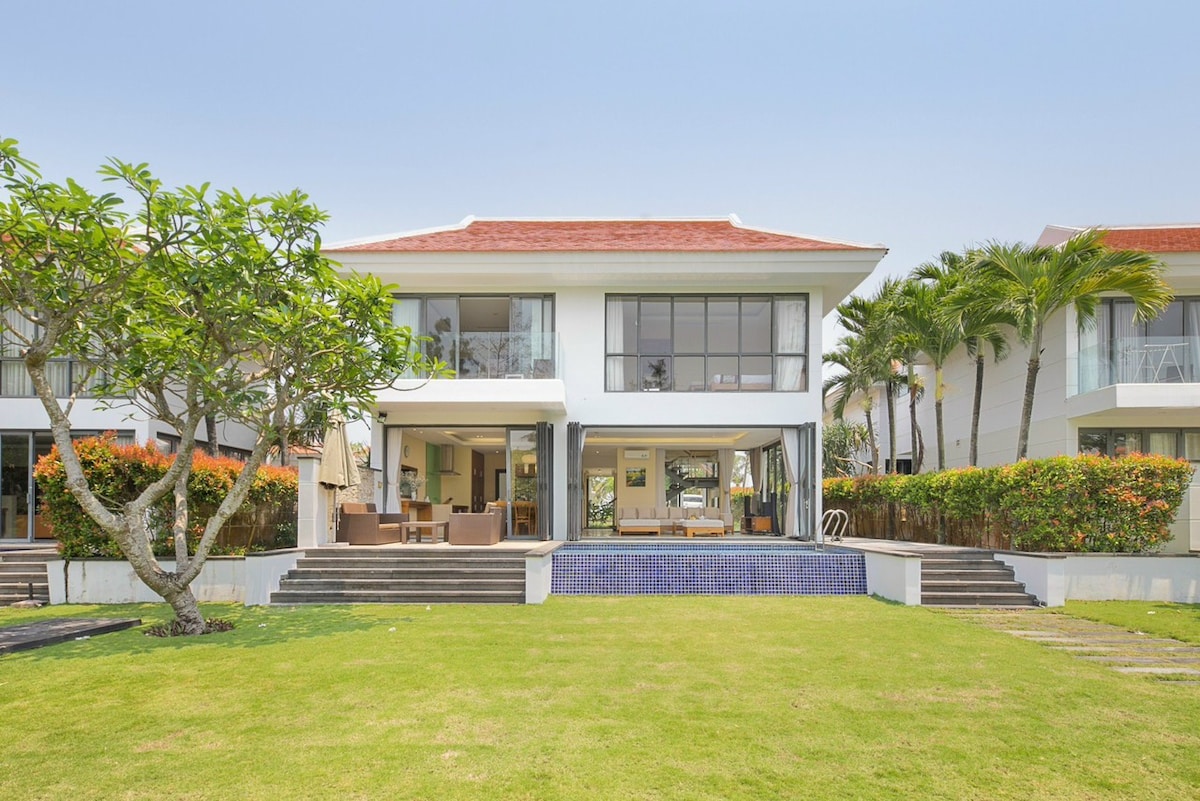 The villa features a spacious landscaped garden surrounded by tropical trees and colorful shrubs. A two-story structure is visible, showcasing large glass windows and an outdoor living area. The private swimming pool is positioned at the base of the steps leading into the garden.