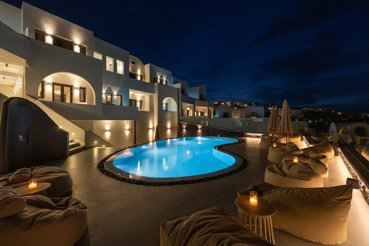 An inviting outdoor area at nighttime features a curved swimming pool illuminated by soft lighting, surrounded by cozy seating options and two candle-lit tables. The elegant architecture of the building is highlighted, with various balconies and decorative elements enhancing the tranquil atmosphere.