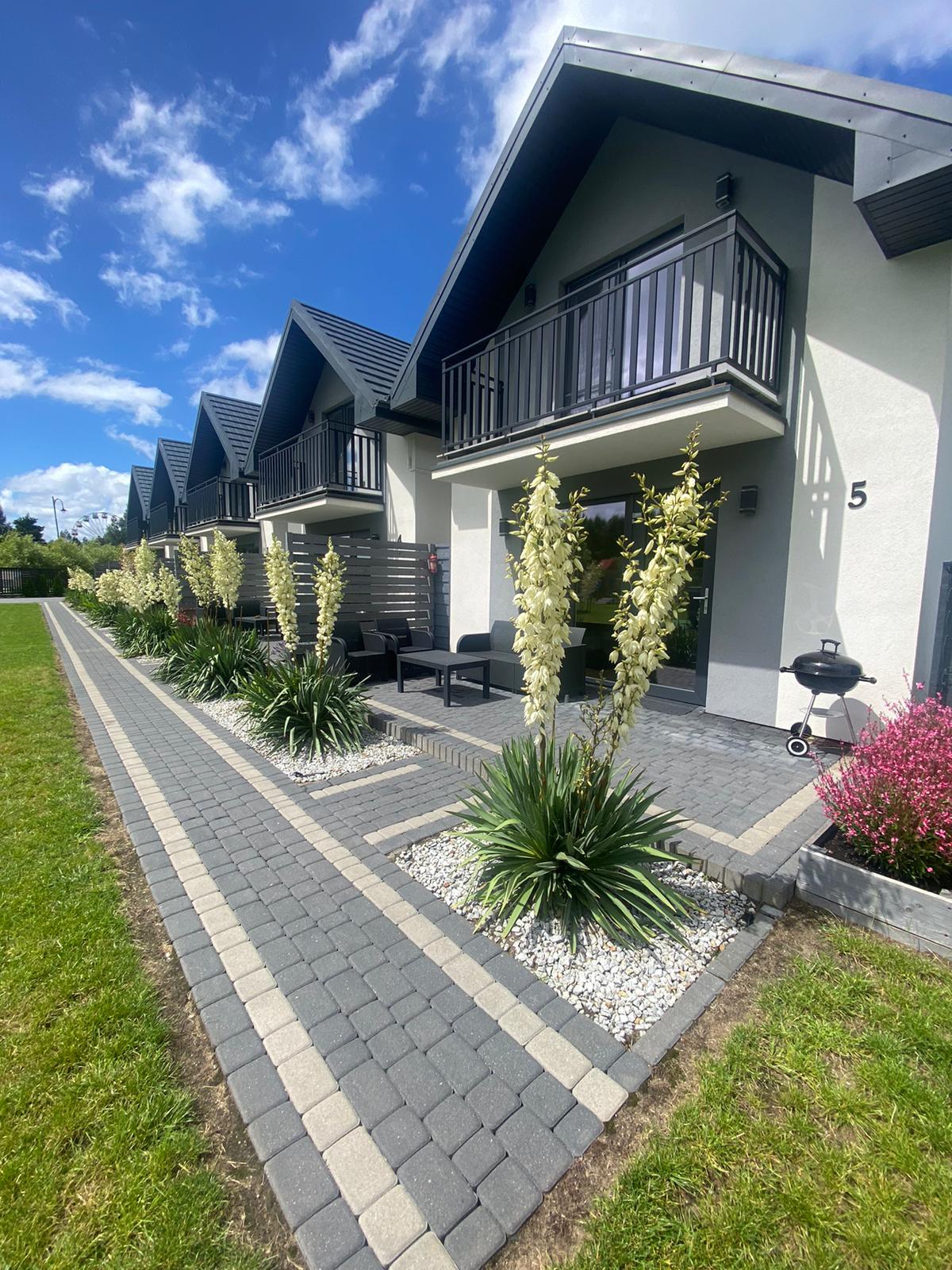 A row of modern accommodations is depicted, featuring balconies with seating arrangements. Well-maintained gardens with lush greenery and flowering plants line the stone pathway leading to the entrance of each unit. A barbecue grill is positioned adjacent to the pathway, enhancing outdoor accessibility.
