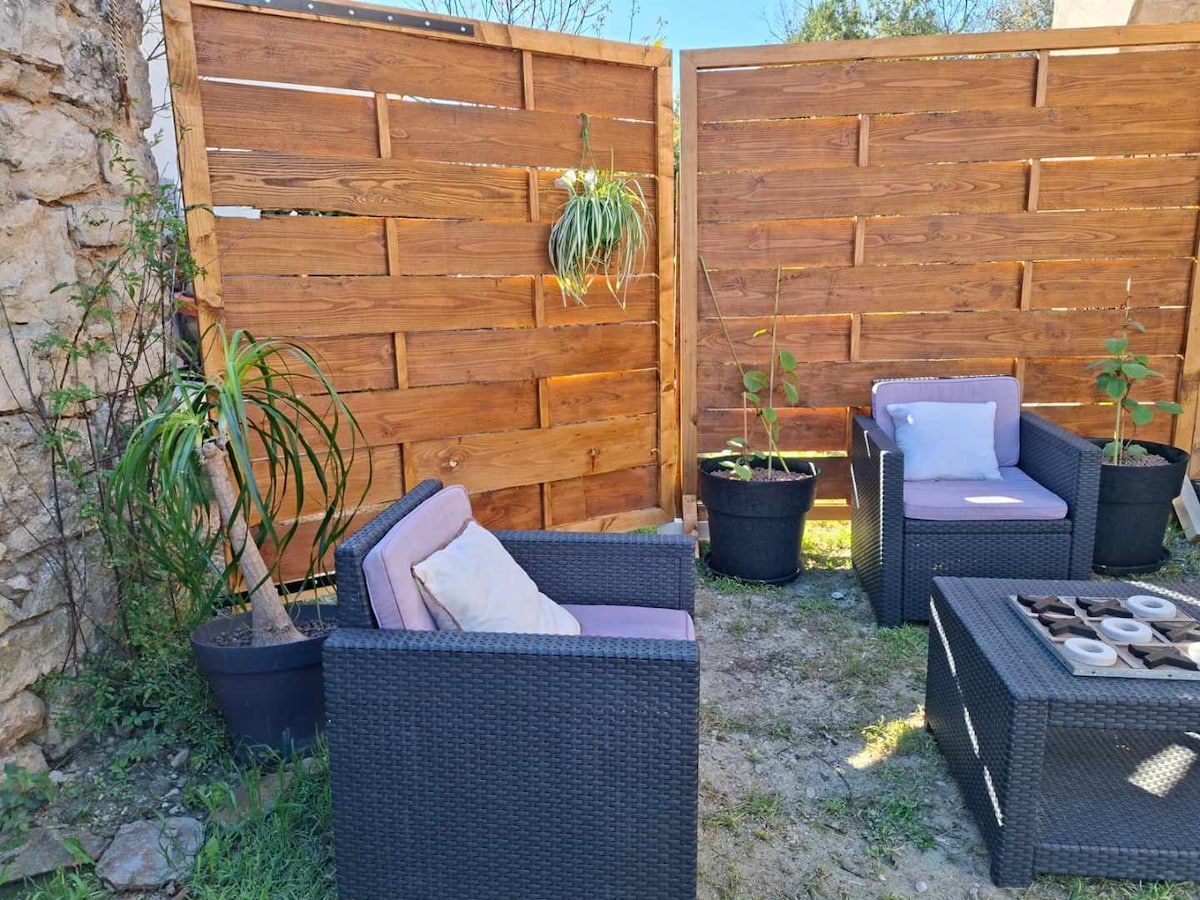 A private garden area features two cozy armchairs and a small fire pit, arranged on a grassy surface. Wooden privacy panels create a secluded space, adorned with potted plants that add a touch of greenery.