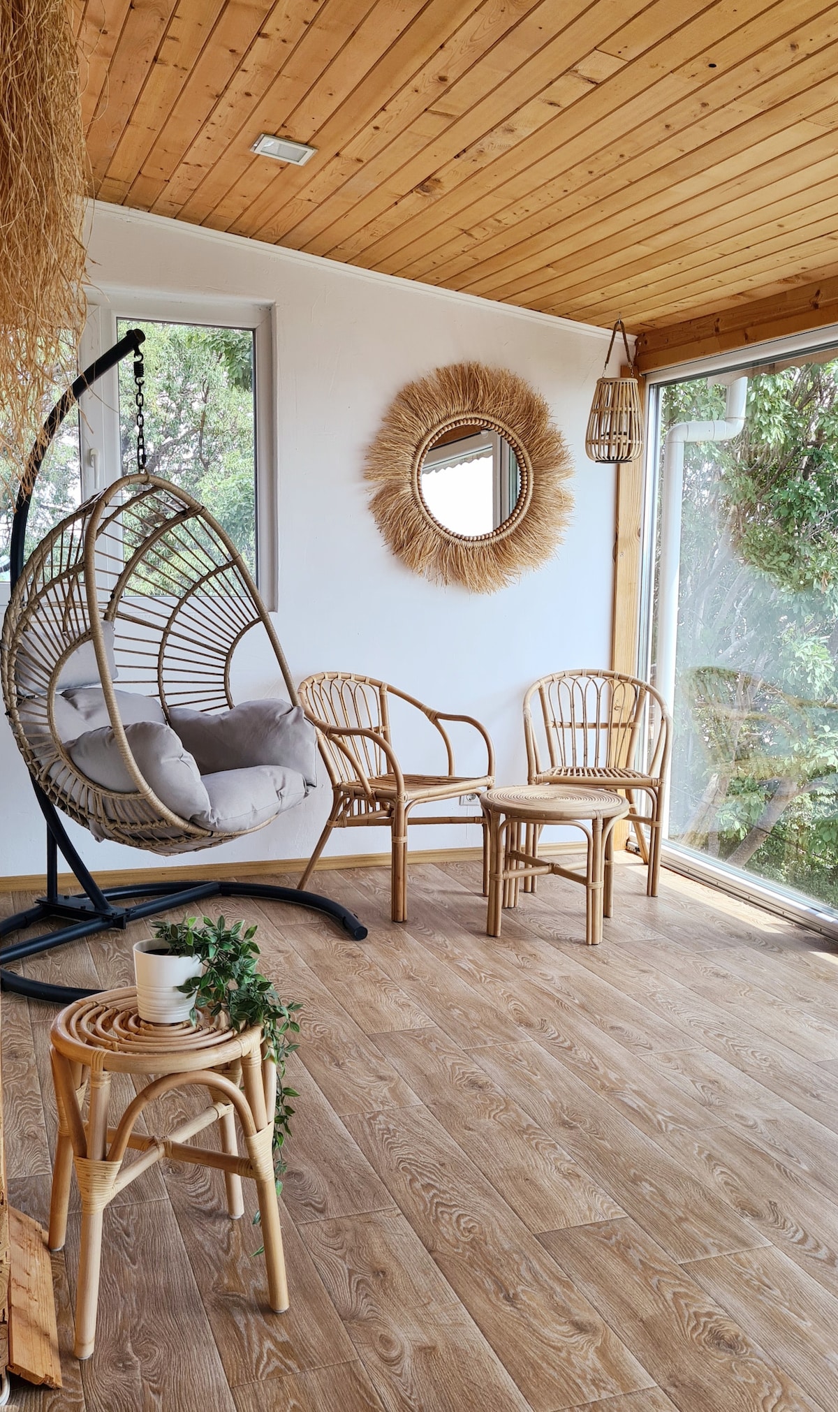 A bright enclosed terrace features a hanging chair and two wicker chairs, creating a comfortable seating area. Natural light fills the space through glass walls. A round mirror and decorative elements enhance the cozy ambiance, while wooden ceiling and flooring add warmth to the setting.