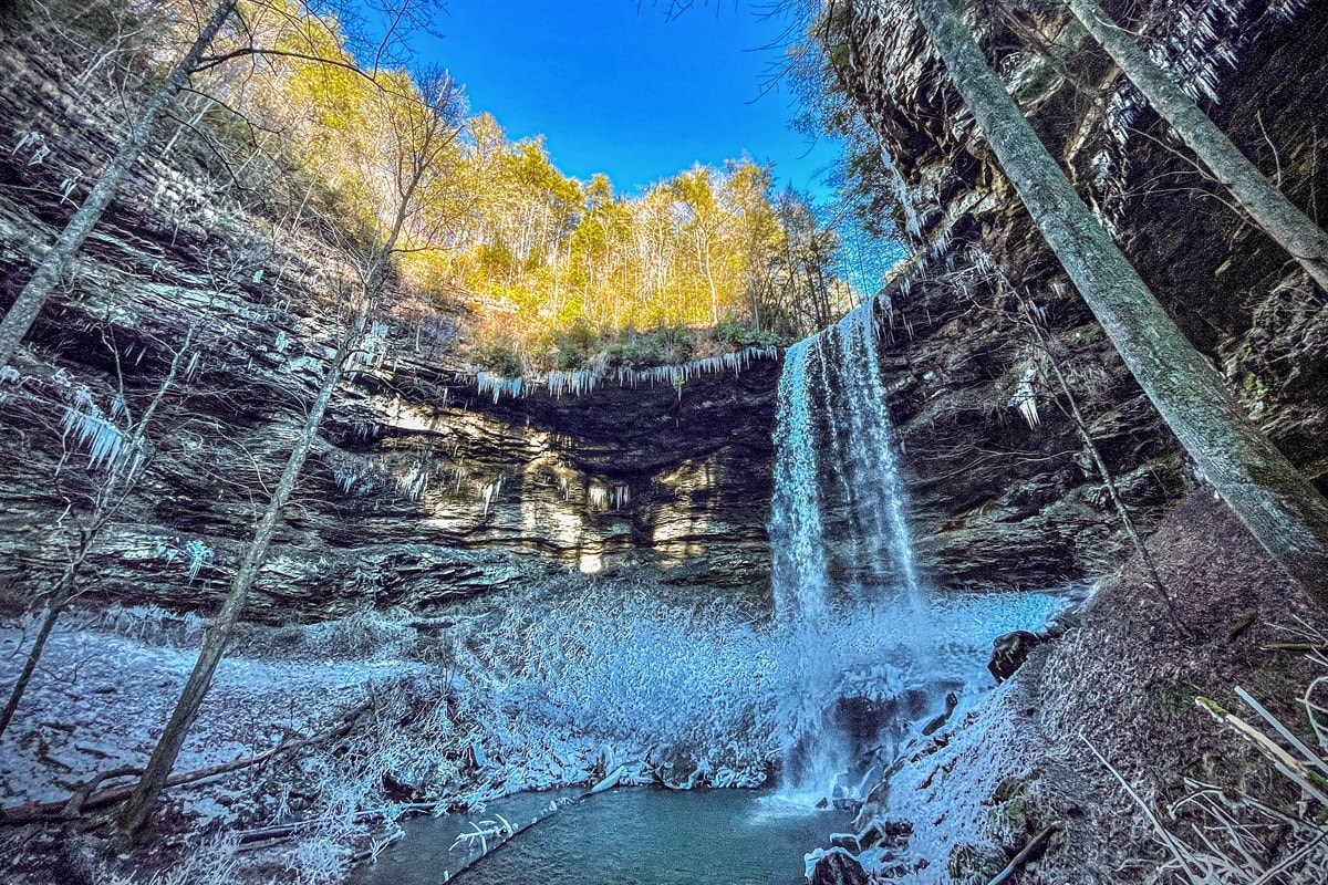 A stunning waterfall cascades down a rocky cliff, surrounded by towering trees. Icicles hang from the ledge above, while patches of snow cover the ground, creating a serene winter landscape. The clear blue sky provides a striking contrast to the natural beauty of the scene.