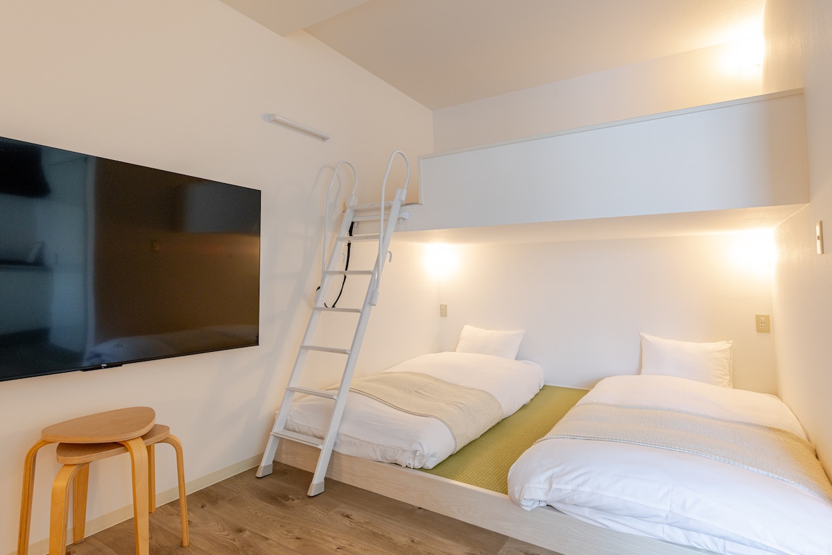 The room features a lofted sleeping area with two spacious beds, complemented by soft white linens. A television is mounted on the wall opposite the beds. A wooden stool adds a touch of warmth to the neutral-toned space, illuminated by soft lighting.