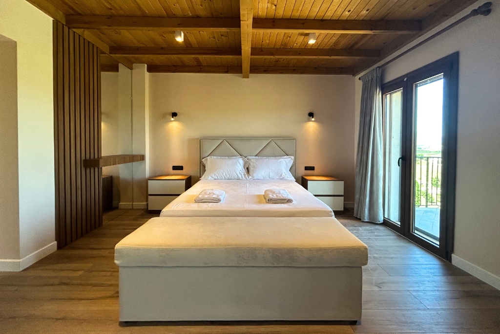 A spacious bedroom features a large bed with neutral bedding set against a wood-paneled ceiling. Soft lighting is provided by wall sconces. Two bedside tables are positioned on either side of the bed, and glass doors open to a bright balcony area.