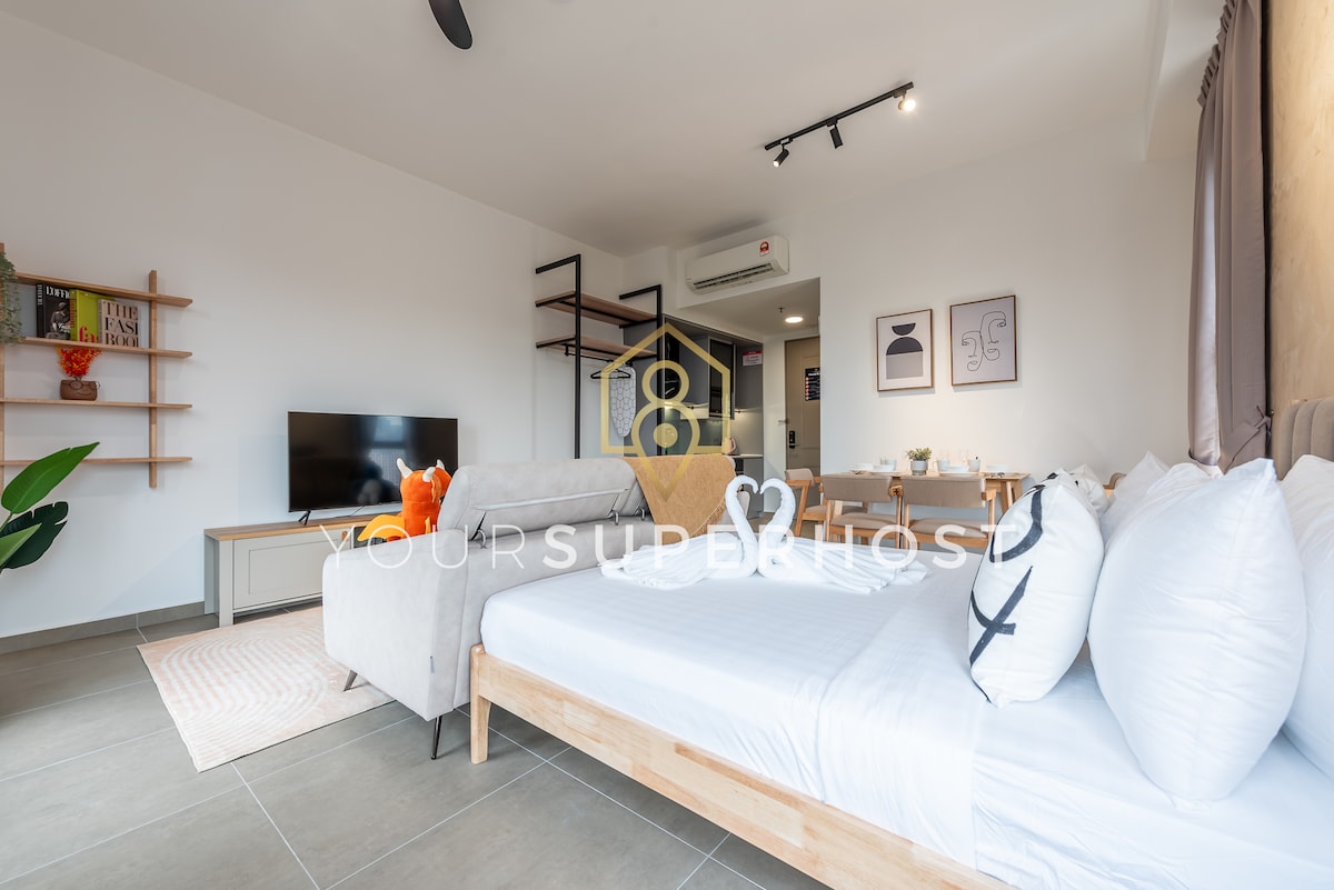 A modern living area features a spacious, light-colored sofa adjacent to a flat-screen TV mounted on the wall. A neatly made bed with fluffy pillows is positioned in the corner. Soft lighting enhances a clean and simple design, complemented by decorative wall art.