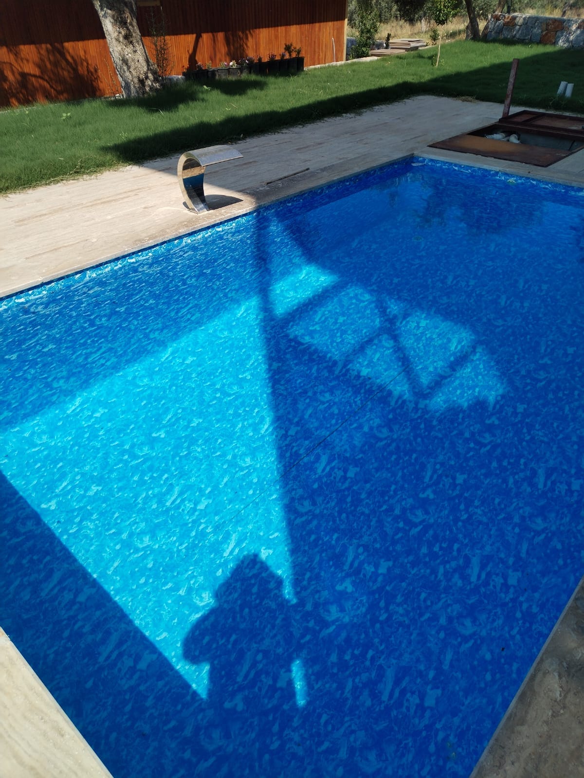 A swimming pool with vibrant blue water is featured, surrounded by wooden decking. The shadow of a tree is cast onto the water, creating a gentle pattern. Lush green grass is visible around the pool area, contributing to a serene outdoor setting.