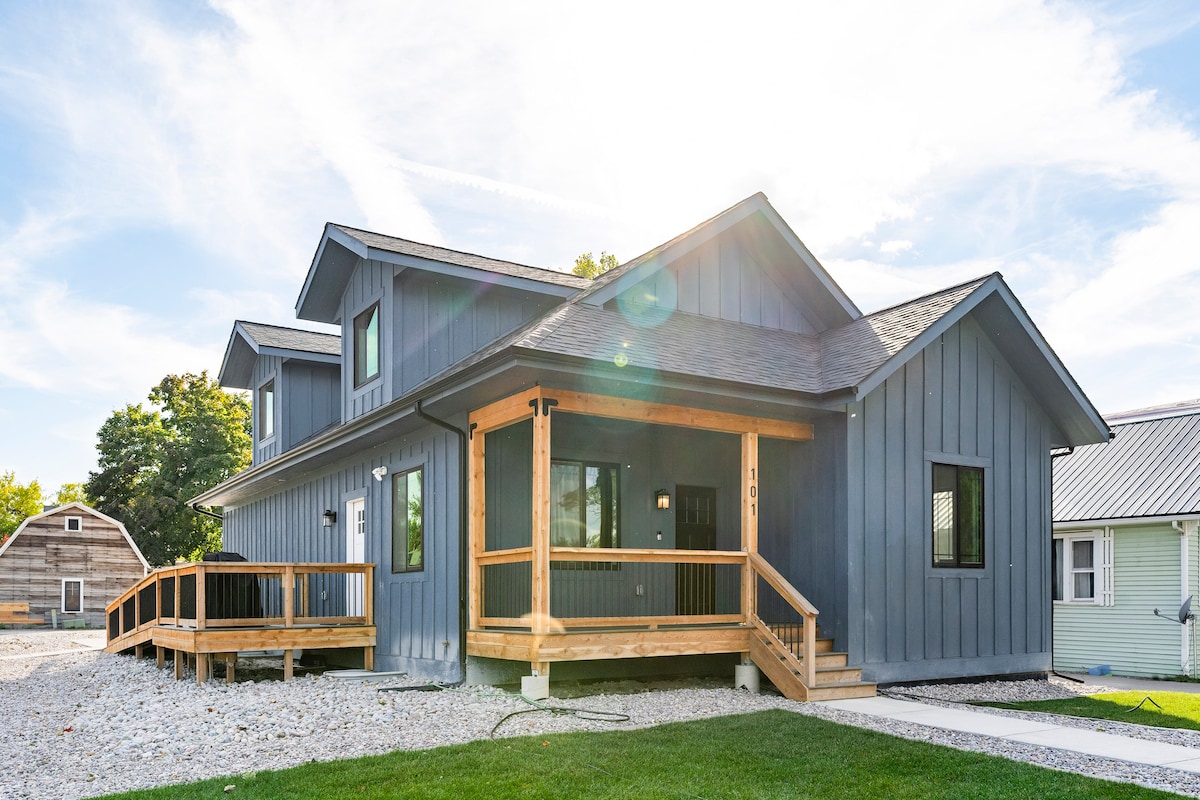 The exterior of a modern two-story home features a covered porch with a wooden railing and a gentle ramp for accessibility. The dark siding is complemented by bright trim, and landscaping consists of gravel and grass patches. Sunlight creates a warm glow against the house.